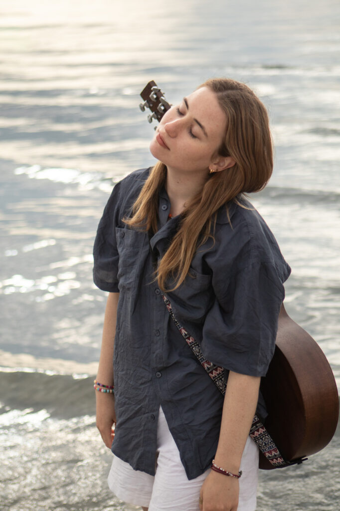 Lina Kahle mit Gitarre auf dem Rücken vor einem Meer.