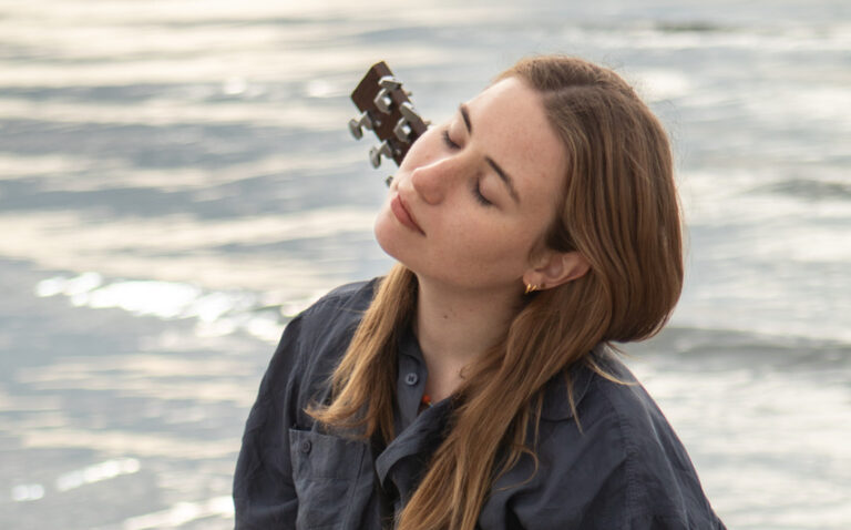 Lina Kahle mit Gitarre auf dem Rücken vor einem Meer.